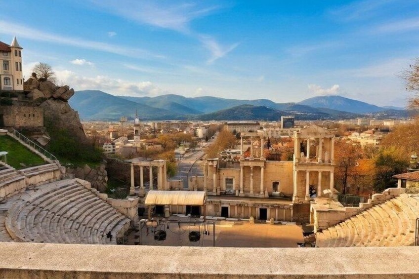 Ancient ruins with columns and a theater, overlooking a city with mountains in the background. 1