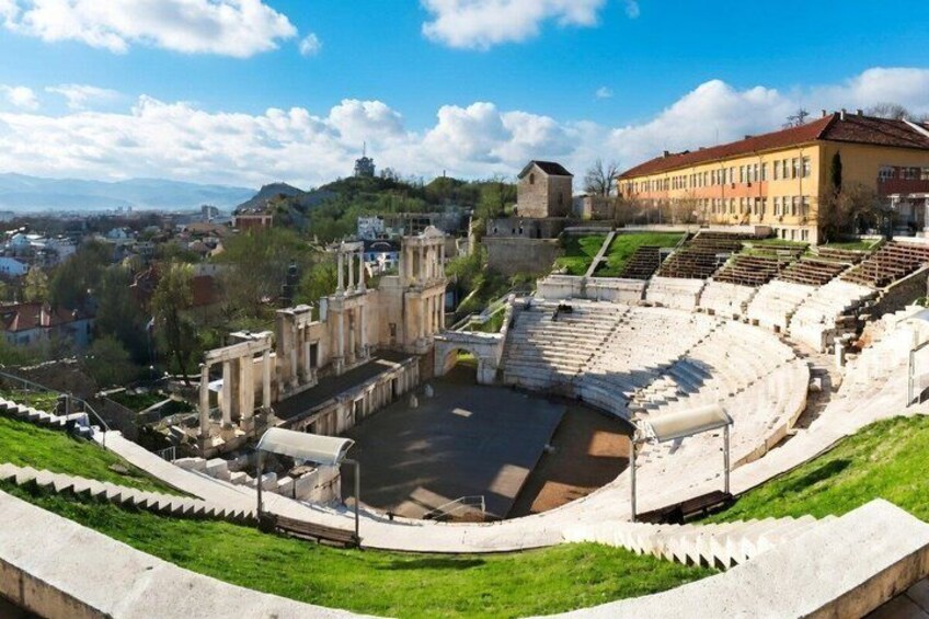 An ancient amphitheater with stone seating and ruins, surrounded by greenery and modern buildings. 2