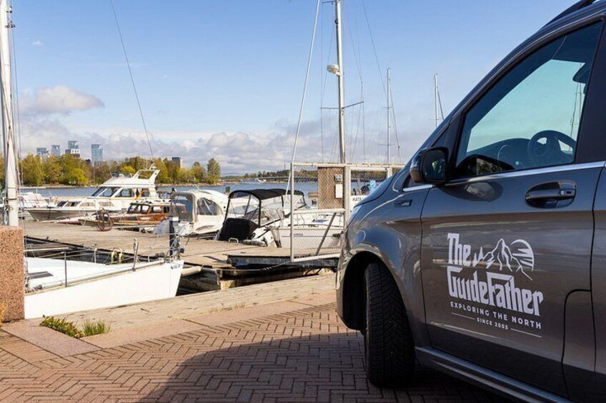 A marina with boats docked, a car with 'The GuideFather' logo, and a city skyline in the background. 2