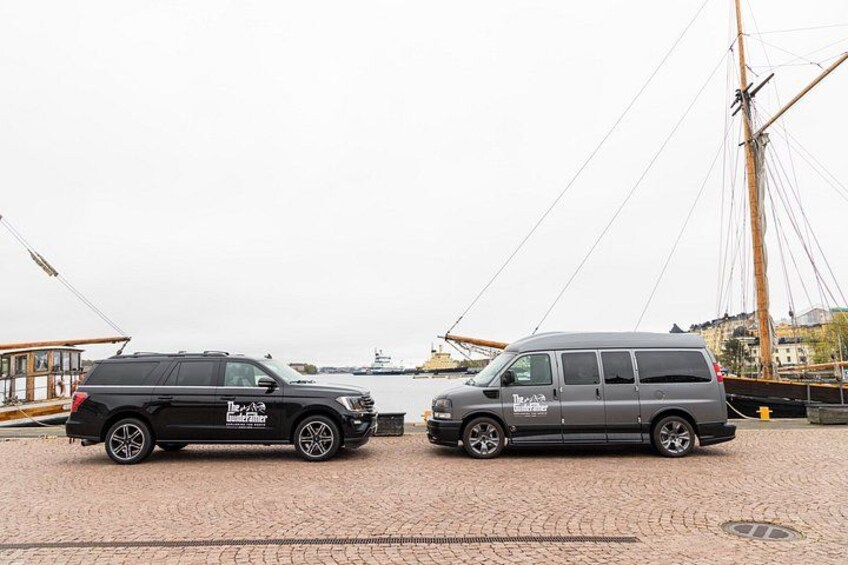 Two vehicles parked on a cobblestone surface with a large ship and a tall mast in the background. 4