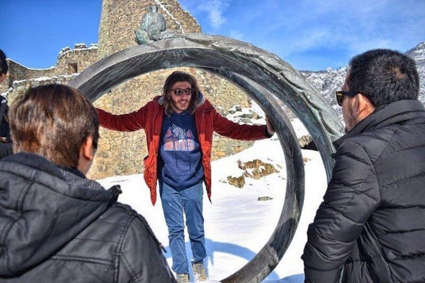 A person standing in a circular frame with arms outstretched, surrounded by snow and rocky terrain. 1
