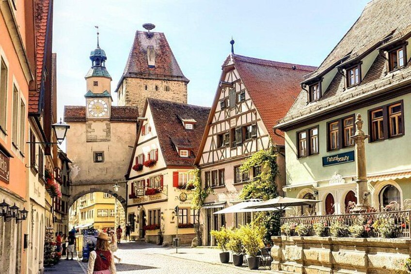 A historic European street with half-timbered buildings, a clock tower, and a fountain. 1