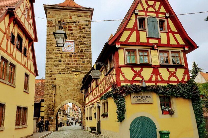 A narrow cobblestone street flanked by traditional half-timbered houses with a stone tower and a clock. 2