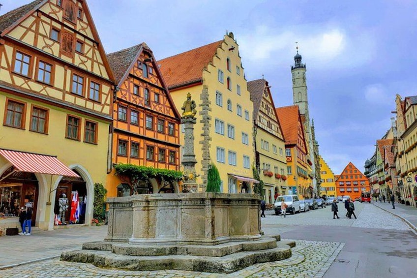 A cobblestone square with historic buildings, a fountain, and a church tower. 3