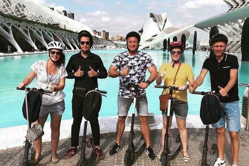 A group of people on hoverboards by a waterfront with modern architecture in the background. 1