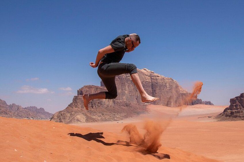 A person jumping in a desert with red sand and rocky formations in the background. 4