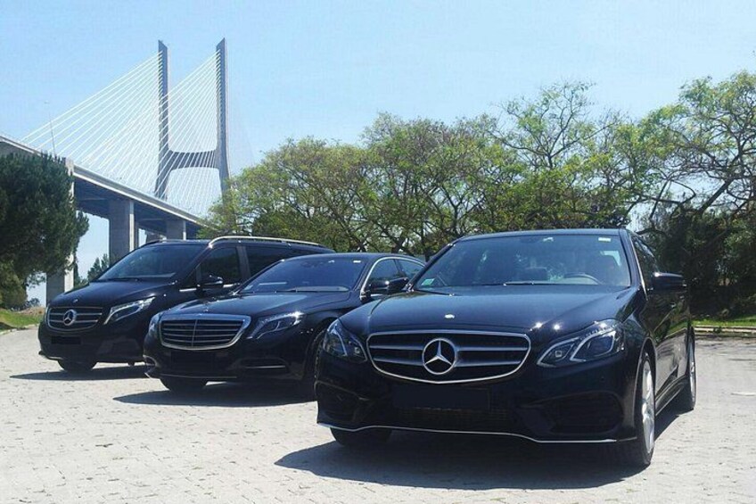 Three black Mercedes-Benz cars parked in a line on a paved surface with a bridge and trees in the background. 2