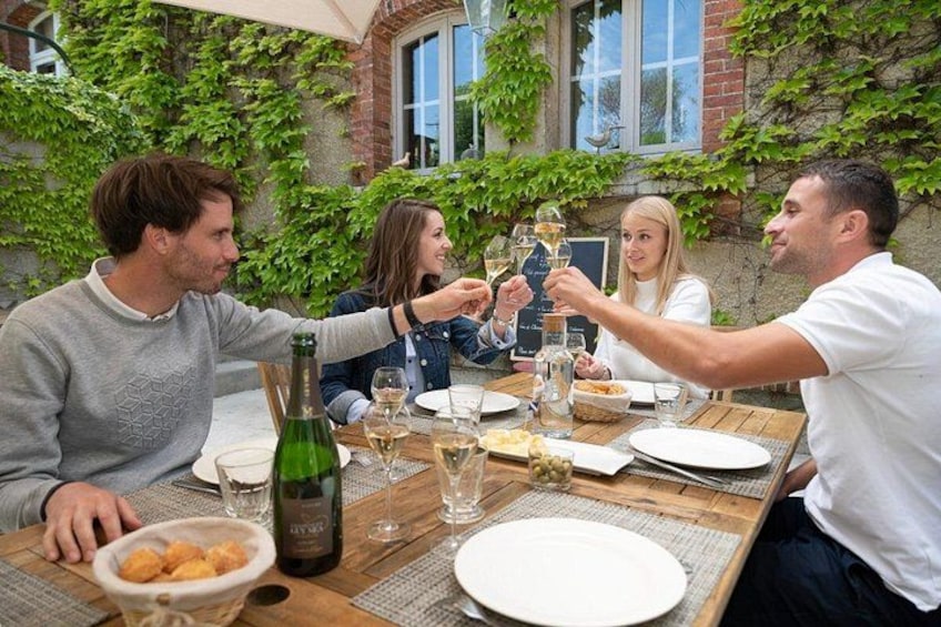 A group of people toasting with wine glasses at a wooden table with a bottle of wine, surrounded by greenery and a brick wall. 1
