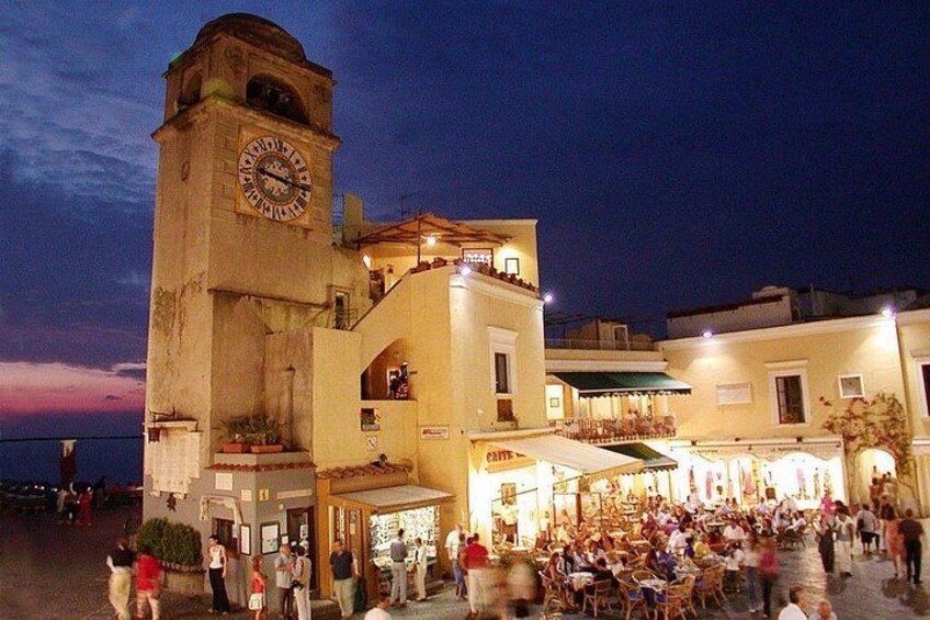 A historic square with a clock tower, outdoor cafes, and people walking around. 2