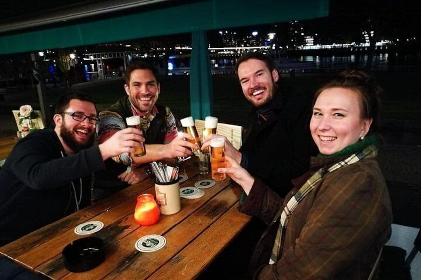 A group of people toasting with beer glasses at a wooden table with coasters, a candle, and a bottle. 2