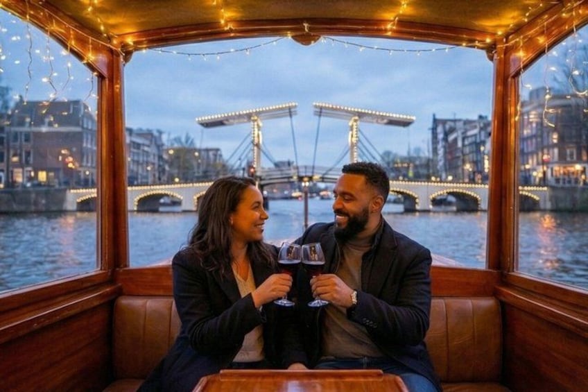 A couple toasting with drinks on a boat with a cityscape and bridges in the background. 2
