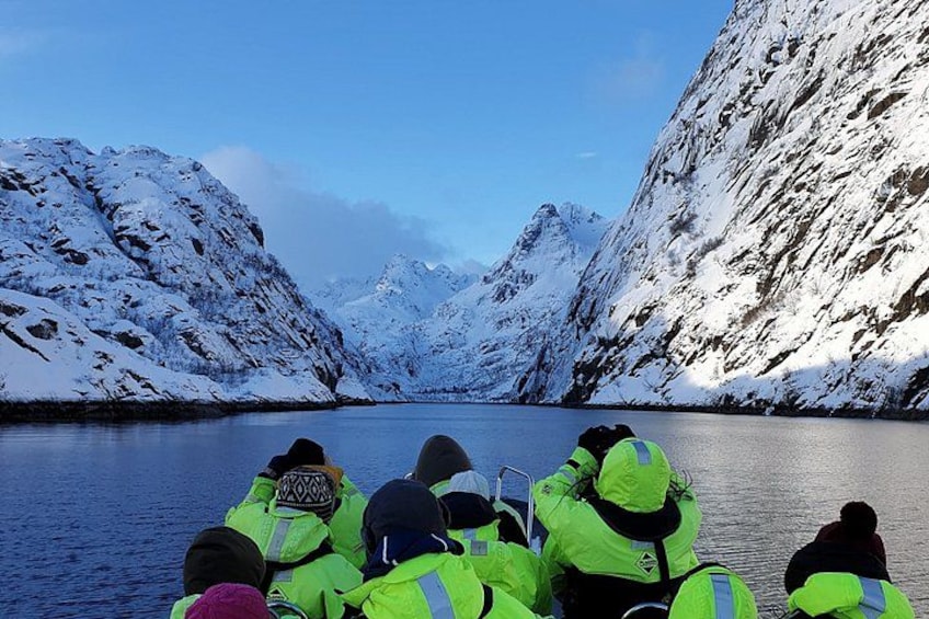 Un gruppo di persone con giacche giallo brillante su una barca, circondato da montagne innevate e un'acqua calma. 1