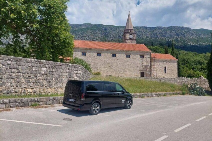 A black van parked in a parking lot with a stone wall, a building with a clock tower, and mountains in the background. 2