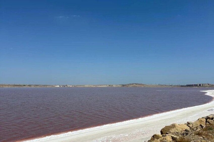 A beach with a red-hued water body, a clear sky, and distant buildings. 2