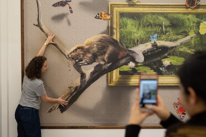Una persona toca un mural grande de un oso en una rama de árbol, con un cuadro enmarcado de un estanque al fondo. 4