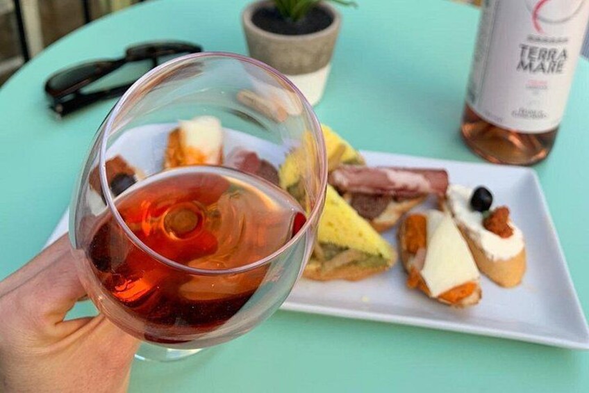 Une main tenant un verre de vin rouge, une assiette avec divers mets et une bouteille de vin Terra Mare sur une table turquoise. 3