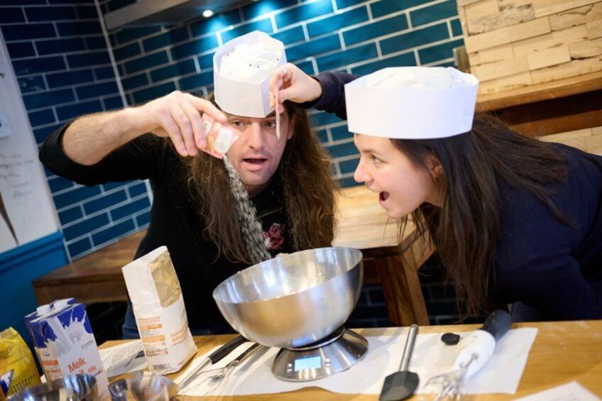 Two people wearing chef hats, a metal mixing bowl, and a wooden table with various food items. 4