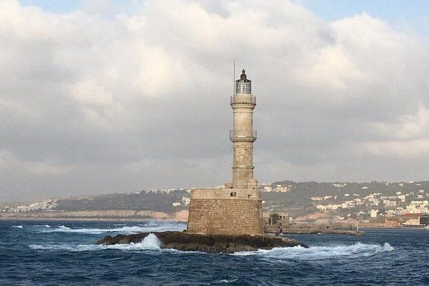 A lighthouse on a rocky outcrop with waves crashing against it, a cloudy sky, and a distant town. 5