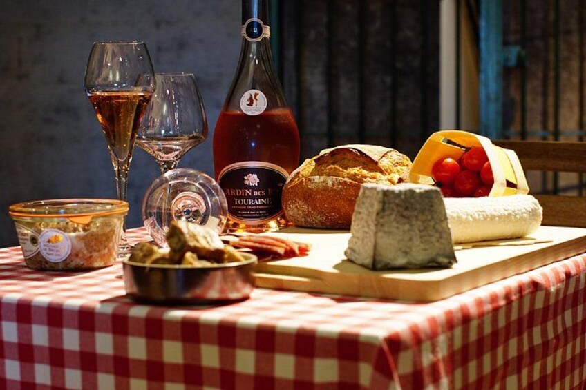 Una mesa con mantel a cuadros rojo y blanco, una botella de vino, copas, una hogaza de pan, un queso redondo, un cuenco de aceitunas y una cesta de tomates. 5