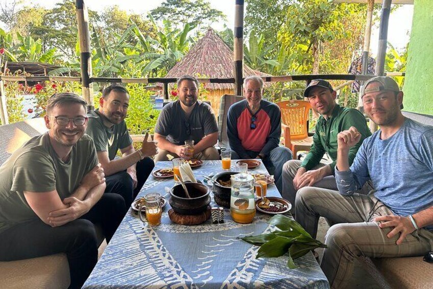 A group of people sitting around a table with food and drinks, outdoors with greenery in the background. 4