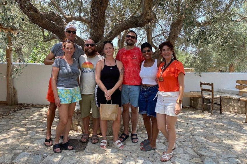 A group of people standing under a tree in a stone-paved area with benches and a white wall in the background. 2