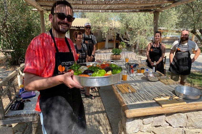 A person holding a large metal tray with various vegetables, a grill, and cooking utensils in an outdoor setting. 4