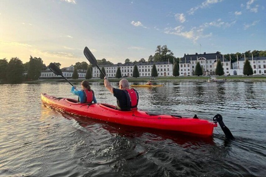 Two people kayaking on a calm lake with a large building in the background. 1