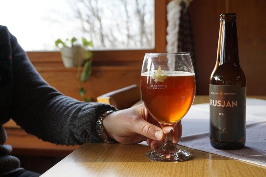 En hand håller ett ölglas bredvid en flaska med etiketten ”RUSJAN IPA” på ett träbord. 5