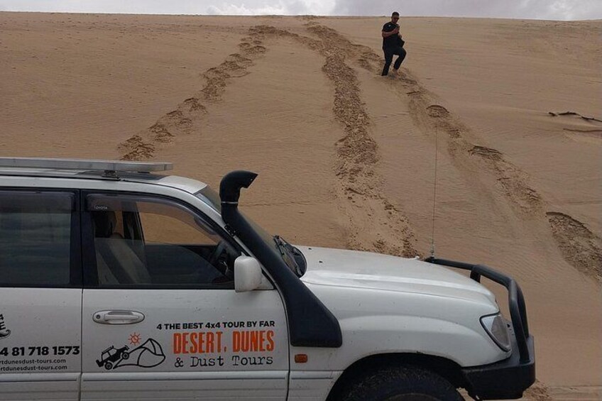 A white vehicle with a desert tour advertisement, a person walking in the sand, and tracks in the sand. 1