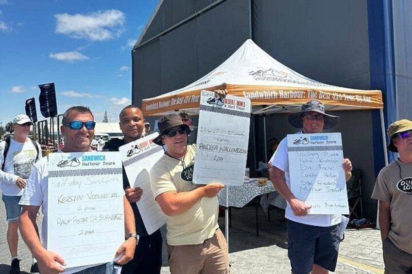 People holding large checkboards with text, standing in front of a tent with a sign that reads 'Sandwich Harbour: The best 24-hour town in the world.' 2