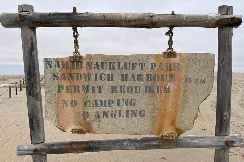 A weathered signpost with text indicating Namib Naukluft Park, Sandwich Harbour, and a permit requirement. 5