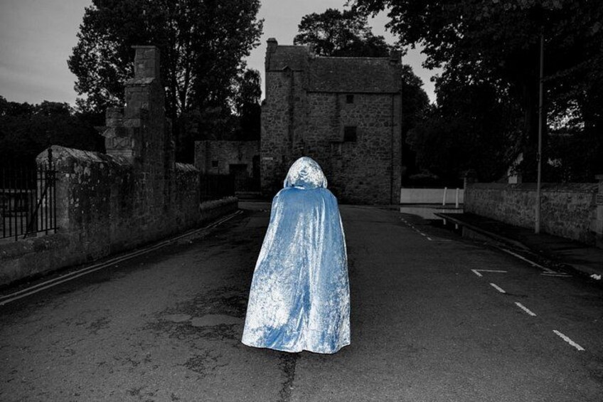 A person in a blue cloak walking down a street with stone buildings and a fence. 5
