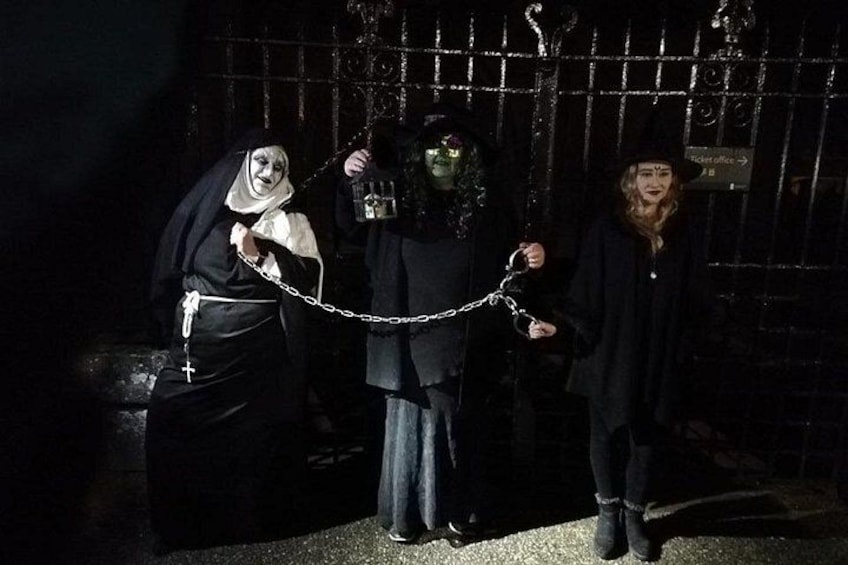 Three individuals in dark clothing, one chained to another, in a dimly lit, barred enclosure. 3