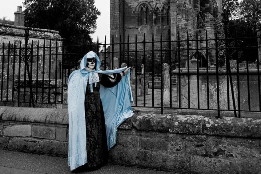 A person in a hooded cloak standing by a cemetery fence. 5