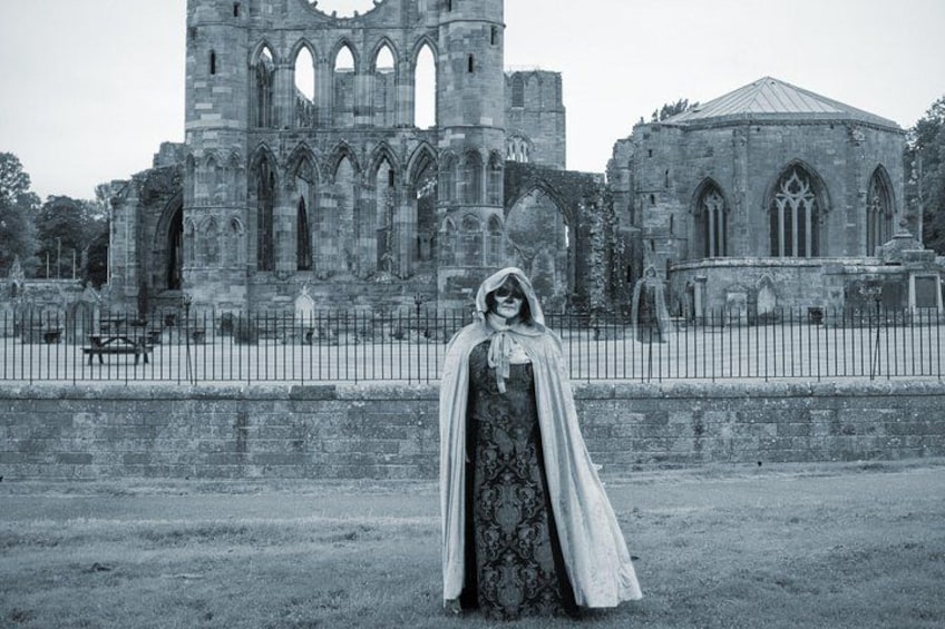 A person in a hooded cloak standing in front of a Gothic-style church with a circular apse and a tall bell tower. 3