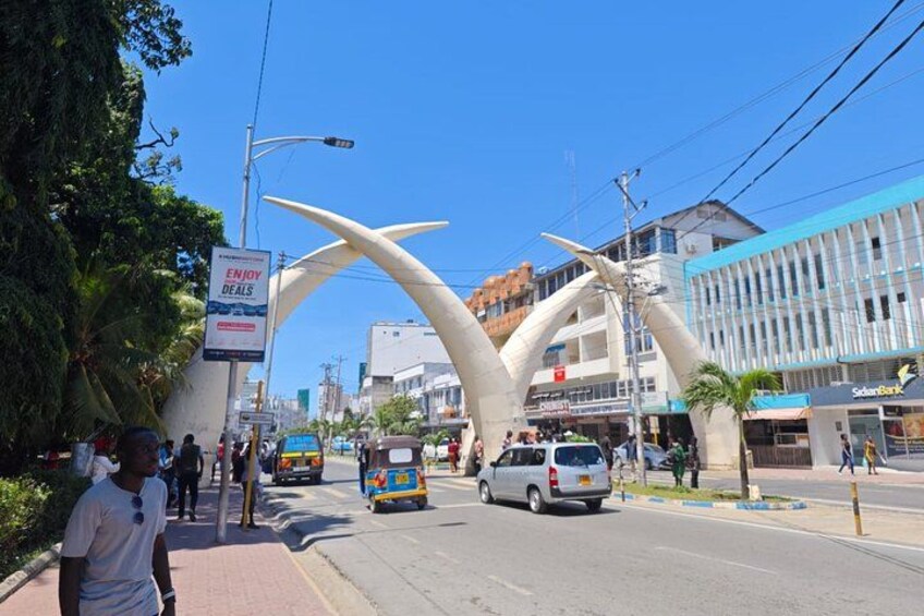 Une rue animée en milieu urbain, caractérisée par une arche blanche distinctive, une enseigne publicitaire annonçant des promotions, ainsi que par la présence de divers véhicules et de piétons. 4
