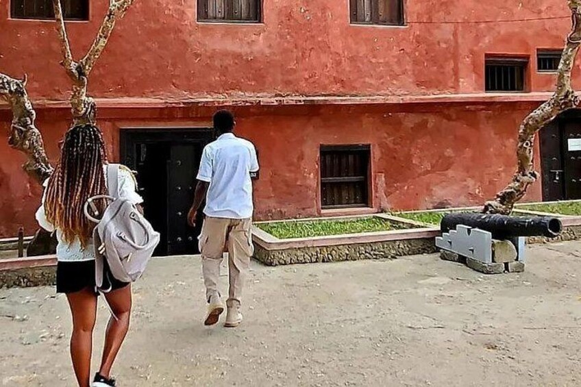 Un bâtiment rouge avec une porte noire, un canon et deux personnes qui marchent. 2