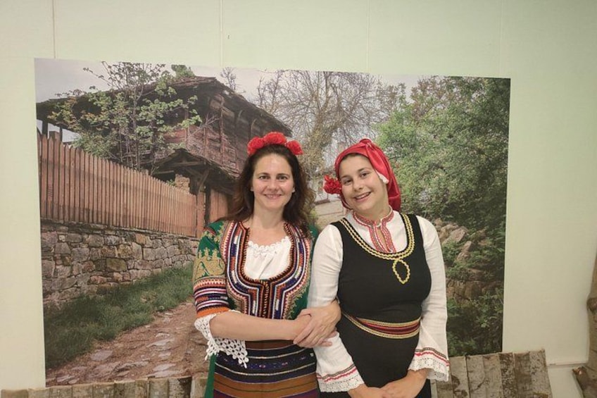 Twee personen in traditionele kleding staan voor een houten gebouw met een stenen muur en een houten hek. 3