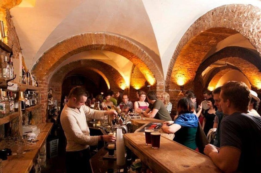 Een bar met bakstenen bogen, een barman die drankjes serveert, en gasten die aan tafels zitten. 1