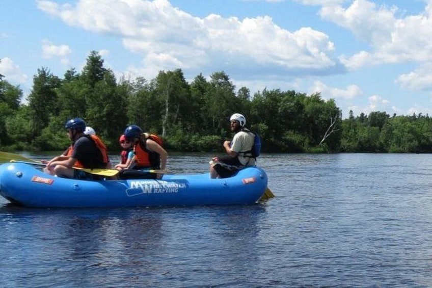 Trois personnes font du rafting sur un radeau gonflable bleu, avec une côte boisée en arrière-plan. 1