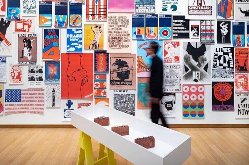 Une galerie où une table blanche expose des objets rouges, le tout étant entouré d’un mur recouvert d’affiches colorées. 2