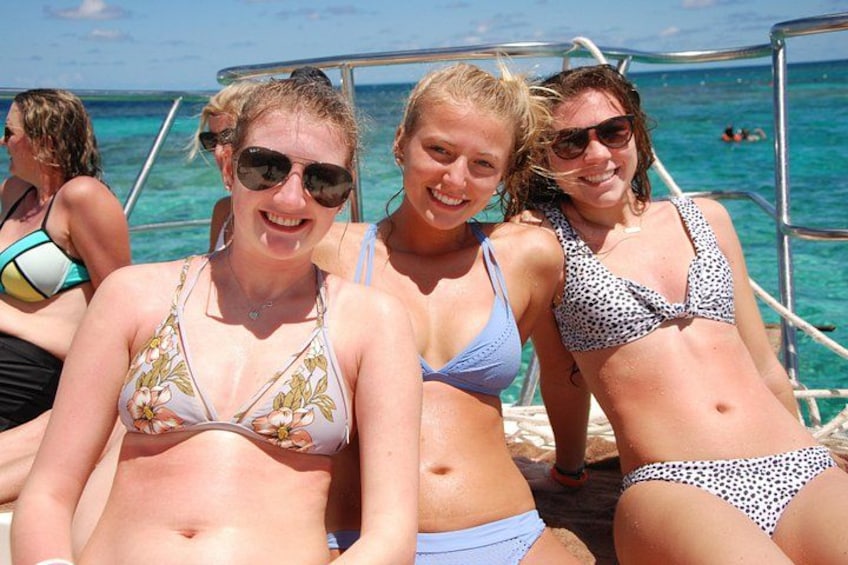 Drie mensen op een boot, in bikini's, met de oceaan en een heldere lucht op de achtergrond. 2