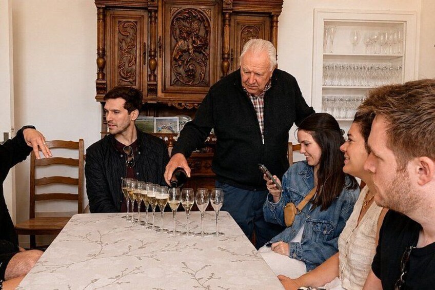 Een groep mensen zit rond een tafel met wijnglazen, een oudere man staat, en een vrouw houdt een telefoon vast. 2