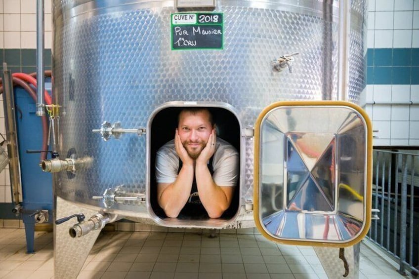 A person inside a stainless steel tank with a door, a sign with 'CUVE N° 2018' and 'Per Manger' on it, and a reflection in a mirror. 4