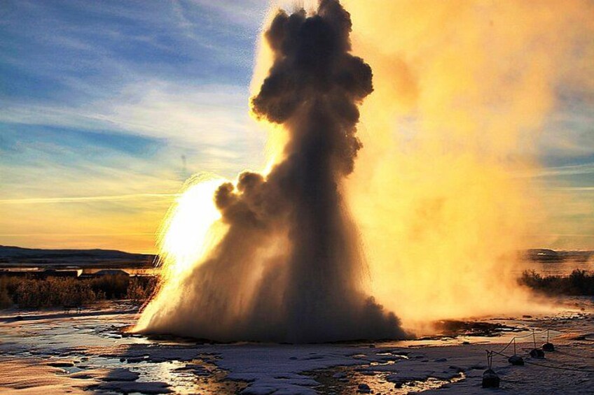 Ein Geysir bricht aus, begleitet von Wasserdampf und Sprühregen vor einem Sonnenuntergangshimmel. 1