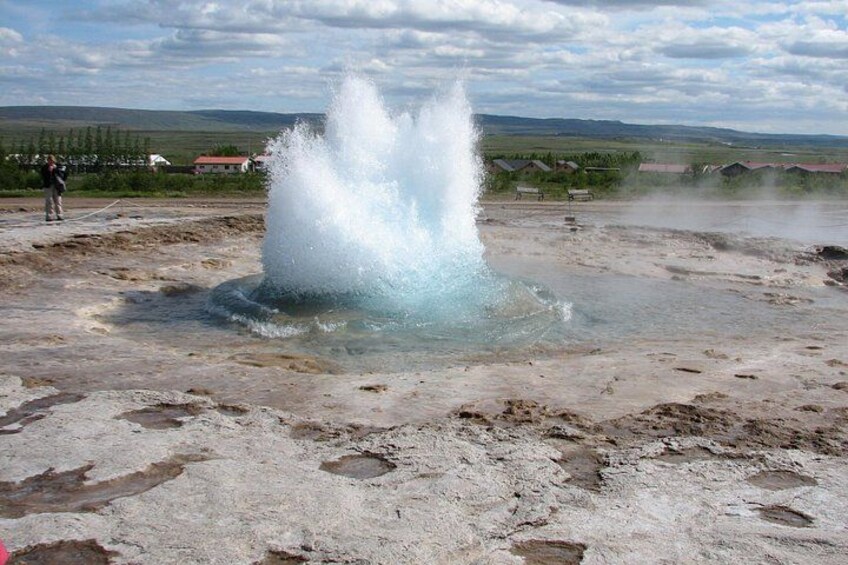Ein Geysir bricht aus und schleudert heißen Dampf in die Luft, umgeben von einer flachen Landschaft mit vereinzelter Vegetation und fernen Gebäuden. 4