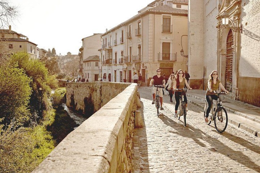 Eine Kopfsteinpflasterstraße mit Radfahrern, einer Steinbrücke und Gebäuden. 2