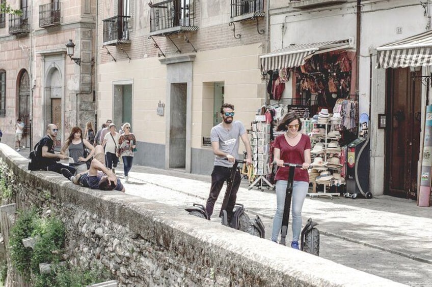 Une rue où l’on voit des gens assis et qui se promènent, un homme sur un scooter, et un magasin de vêtements et de chaussures. 2
