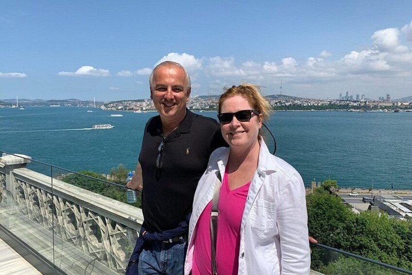 Two people standing on a balcony overlooking a body of water with a city skyline in the background. 2