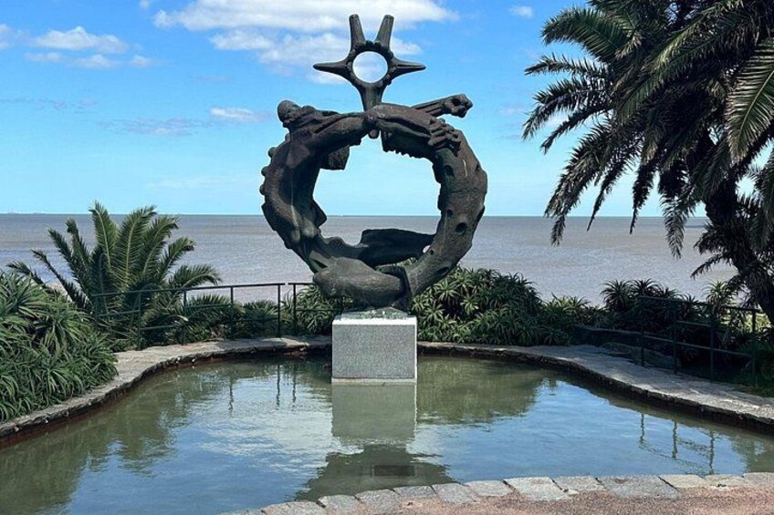 En skulptur af en hånd der holder en stjerne, omgivet af et vandbassin, med en strand og palmer i baggrunden. 3
